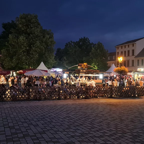 Eine belebte Veranstaltung unter freiem Himmel, mit zahlreichen Menschen, die an Ständen essen und trinken. Über dem Platz herrscht nächtliche Stimmung, erleuchtet von Lampen und einem dunklen Himmel. Verschiedene Essensstände und ein festlicher Rahmen sind sichtbar.