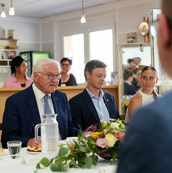 Eine Gruppe von vier Personen sitzt an einem Tisch. Ein älterer Mann mit Brille und Anzug blickt nachdenklich, während zwei jüngere Männer und eine Frau aufmerksam zuhören. Auf dem Tisch befinden sich Essen und Blumenarrangements.