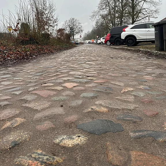 Eine klare Sicht auf eine gepflasterte Straße, die in der Ferne verläuft. Rechts sind geparkte Autos zu sehen, während die Umgebung von kahlen Bäumen und einem grauen Himmel geprägt ist. Die Stimmung wirkt ruhig und nüchtern.