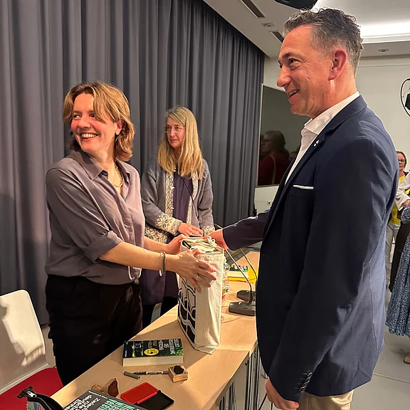 Bei einer Veranstaltung in Innenraum reicht eine lächelnde Frau einem elegant gekleideten Mann über einen Tisch eine verpackte Tasche. Auf dem Tisch liegen Bücher und Gegenstände; die Szene wirkt freundlich und ungezwungen.