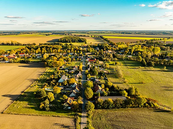 Luftaufnahme eines kleinen Dorfes umgeben von Feldern und Wiesen in der herbstlichen Landschaft. Die Häuser sind von Bäumen und Sträuchern umgeben, die Farben der Blätter variieren zwischen Grün und Gelb. Im Hintergrund erstrecken sich sanfte Hügel und weite Felder.