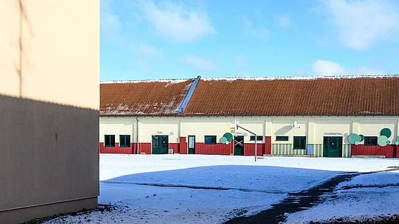 Eine schneebedeckte Fläche vor einem Gebäude mit farbigen Fenstern und einem roten unteren Bereich. Auf der linken Seite befindet sich eine Wand, während auf der rechten Seite das Gebäude und ein Basketballkorb sichtbar sind. Der Himmel ist blau mit vereinzelten Wolken.