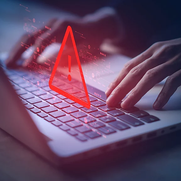 Hände tippen auf einer Laptop-Tastatur, darüber erscheint ein rotes Warndreieck mit Ausrufezeichen – Hinweis auf Sicherheitsproblem oder Systemfehler.