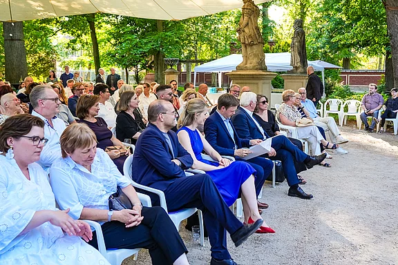 Eine Gruppe von Menschen sitzt in einem Park unter einem Sonnenschutz. Die Anwesenden sind in formeller Kleidung und scheinen einer Veranstaltung zuzuhören. Im Hintergrund sind Bäume und Statuen sichtbar, was eine festliche Atmosphäre schafft.