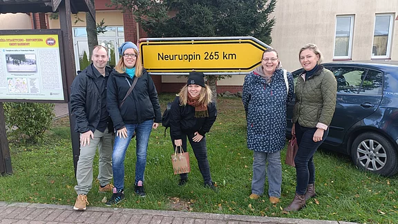 Eine Gruppe von fünf Personen steht lächelnd vor einem gelben Schild, das "Neuruppin 265 km" anzeigt. Die Personen tragen verschiedene Winterjacken und sind in einer grünen Umgebung. Im Hintergrund ist ein Auto und ein informatives Schild sichtbar.