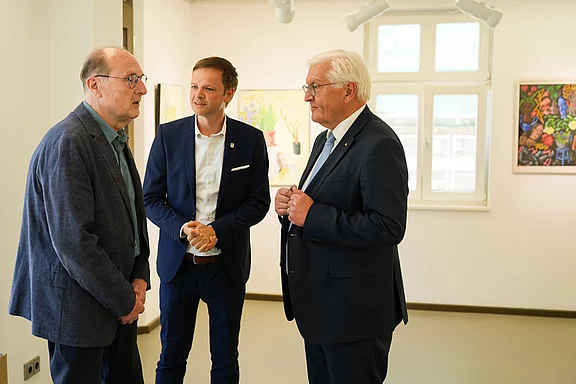 Drei Männer stehen in einer Galerie und unterhalten sich. Einer trägt eine Brille und hat ein graues Jackett an, der zweite trägt einen hellen Anzug und der dritte ist elegant gekleidet. Im Hintergrund sind farbenfrohe Kunstwerke an den Wänden zu sehen.