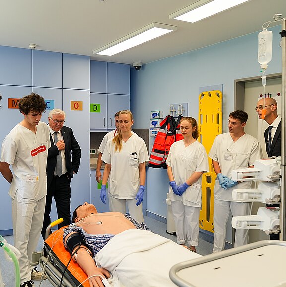 Eine Gruppe von fünf Personen, darunter Medizinstudenten und Fachkräfte, steht um einen Patienten, der auf einer Liege liegt. Sie beobachten eine medizinische Behandlung in einem modernen Klinikraum. Der Patient zeigt eine entspannte Haltung, während die Fachkräfte konzentriert agieren.