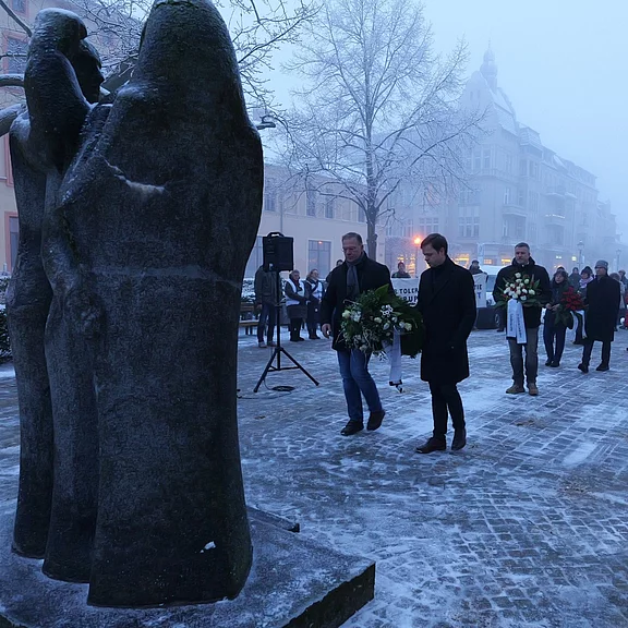 In einer winterlichen Stadtlandschaft gehen Trauernde mit Kränzen zu einem steinernen Denkmal. Der Boden ist vereist, Nebel liegt in der Luft. Menschen stehen respektvoll beisammen, offenbar bei einer Gedenkveranstaltung.