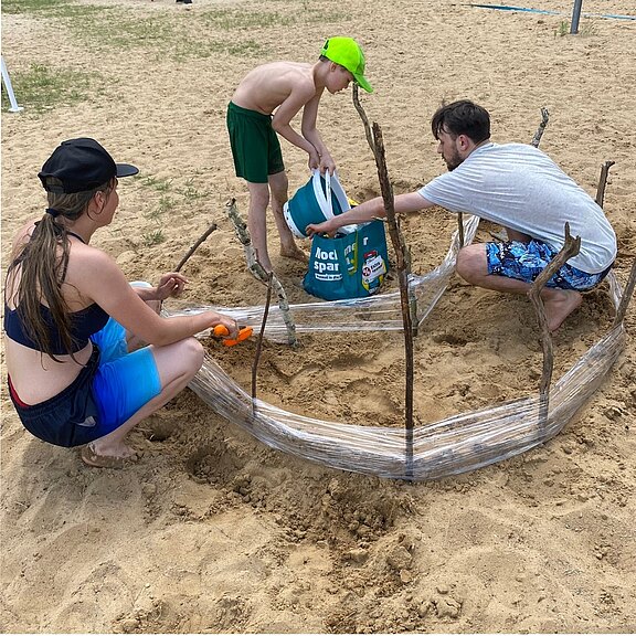 Am Strand bauen ein Kind und zwei Erwachsene gemeinsam einen runden Sandbau: Stöcke und transparente Folie formen eine niedrige Umrandung, daneben ein Eimer mit Material.