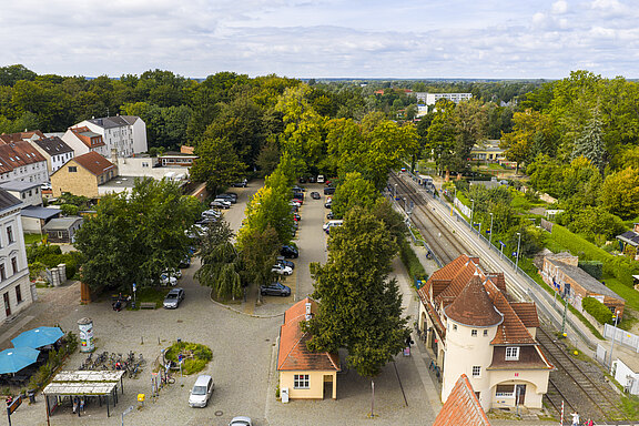 Luftaufnahme des Bahnhofes Neuruppin Rheinsberger Tor, rechts verlaufen Gleise neben einem historischen Stationsgebäude mit roten Ziegeldächern, links ein Parkplatz mit zahlreichen Autos und ein Platz mit Fahrrädern und wenigen Passanten, umgeben von der Stadtmauer, dichten Bäumen und historisch gestalteten Wohnhäusern.