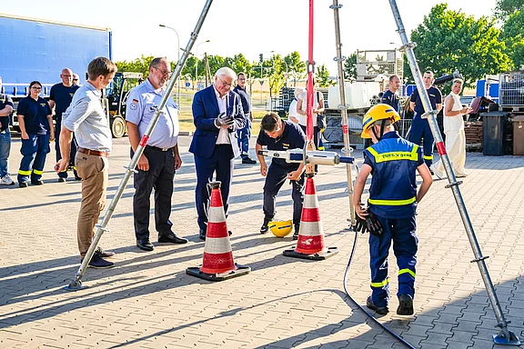 Eine Gruppe von Personen, darunter Feuerwehrleute und offizielle Vertreter, beobachtet einen jungen Feuerwehrmann, der ein Übungsgerät bedient. Warnmarkierungen und eine Sicherheitsvorrichtung sind sichtbar. Die Szene findet im Freien, auf einem Platz mit gepflastertem Boden statt.