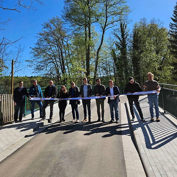 Zehn Personen eröffnen symbolisch eine neue Brücke: Sie stehen nebeneinander und halten ein blau-weißes Band quer über den Weg, das in der Mitte durchschnitten wird. Heller Sonnentag, Bäume im Hintergrund.