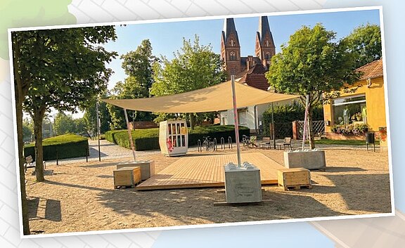 Holzdeck mit großem Sonnensegel auf einem Platz, umgeben von Bäumen und Sitzgelegenheiten; daneben ein kleiner Kiosk. Ruhige, sonnige Parkszene.