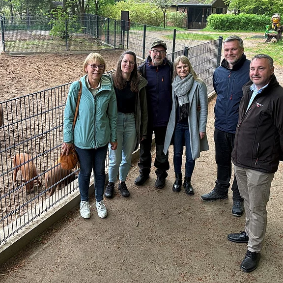 Sechs Erwachsene stehen nebeneinander vor dem Zaun eines Tiergeheges in einem Park und lächeln in die Kamera. Im Gehege wühlt ein kleines braunes Wildschwein im Sand. Grüne Bäume und weitere Gehege im Hintergrund.