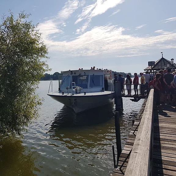 Menschen warten auf einem hölzernen Steg, um in ein kleines weißes Fahrgastboot einzusteigen, das am Ufer eines ruhigen Sees anlegt. Sonniger Tag mit leicht bewölktem Himmel.