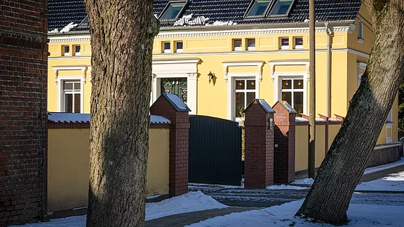 Ein gelbes Gebäude mit weißen Fenstern und einem geschmückten Eingang ist durch eine schwarze Toröffnung sichtbar. Rundherum sind Schnee und Bäume zu sehen, die eine winterliche Atmosphäre schaffen. Der Eingang wird von roten Backsteinpfeilern flankiert.