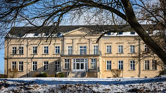 Ein beeindruckendes, klassisches Gebäude mit cremefarbener Fassade und vielen Fenstern. Vor dem Gebäude befindet sich eine Treppe, die zu einem Balkon führt. Der Boden ist mit Schnee bedeckt und im Hintergrund sind kahle Bäume zu sehen. Der Himmel ist blau und klar.