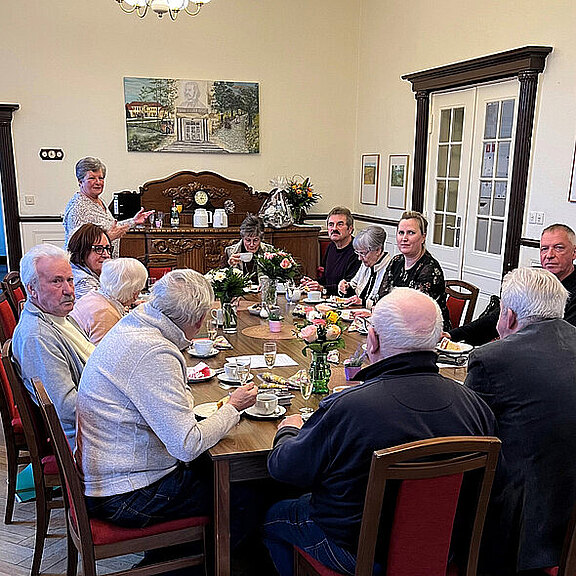 Eine Gruppe von zwölf Personen sitzt an einem runden Tisch, der mit Geschirr und Blumen dekoriert ist. Einige genießen Kaffee und Kuchen. Eine Person steht am Tisch und serviert. Der Raum ist hell und hat eine freundliche Atmosphäre.