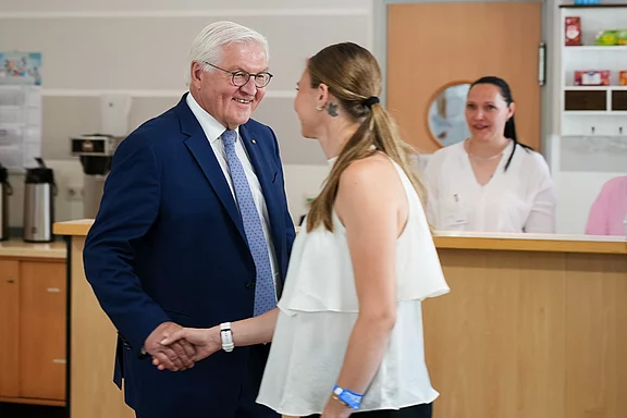 Der deutsche Bundespräsident begrüßt eine junge Frau mit einem Händedruck. Beide lächeln freundlich. Im Hintergrund steht eine weitere Frau, die den Moment beobachtet. Die Szene findet in einer hellen Innenraumumgebung statt.