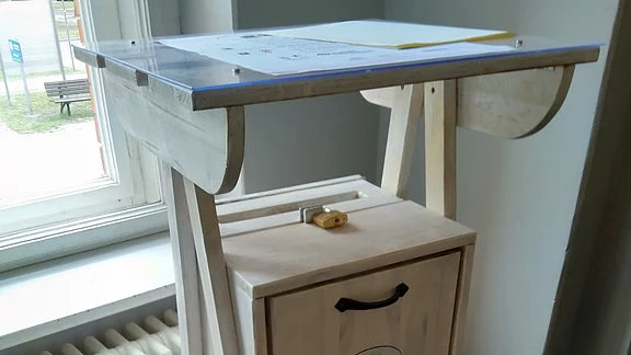 Hohe hölzerne Sammelbox mit integriertem Stehpult. Oben eine flache Schreibfläche mit Dokument unter Plexi; darunter ein verschließbarer Kasten mit Griff und Etikett „Post“. Dient zum Einwerfen von Nachrichten oder Vorschlägen.