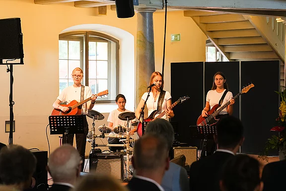 Eine vierköpfige Band spielt live in einem Raum. Die Musikerinnen stehen auf einer Bühne und spielen verschiedene Instrumente, darunter Gitarre und Schlagzeug. Im Vordergrund sind Zuhörer zu sehen, die der Darbietung aufmerksam lauschen.