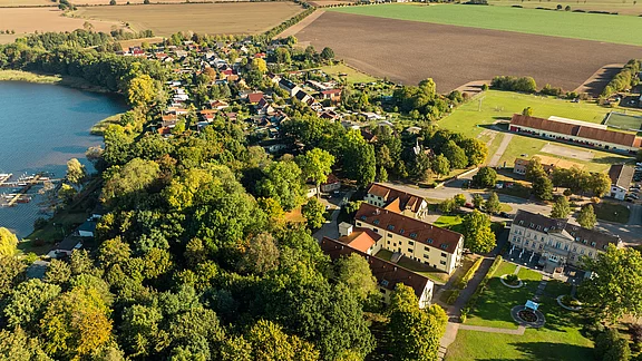 Luftaufnahme eines ländlichen Gebiets mit einem ruhigen See, umgeben von Bäumen und grünen Feldern. Im Vordergrund sind einige Gebäude und ein kleiner Park zu sehen, während sich im Hintergrund die Siedlung mit mehreren Häusern erstreckt.