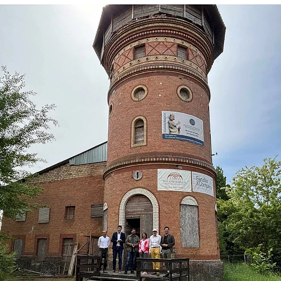 Historischer runder Backsteinturm, teils mit Brettern vernagelt. Ein Plakat hängt an der Fassade. Auf einer kleinen Holzplattform davor stehen sechs Personen, offenbar zu einer Besichtigung. Bäume rahmen das Gebäude.