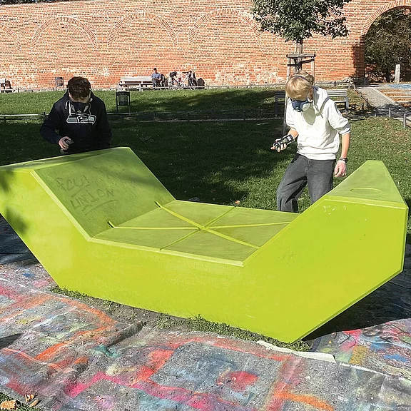 Zwei Personen betrachten und fotografieren eine große, knallgrüne, geometrische Parkbank oder Skulptur auf bemaltem Beton in einer Grünanlage.