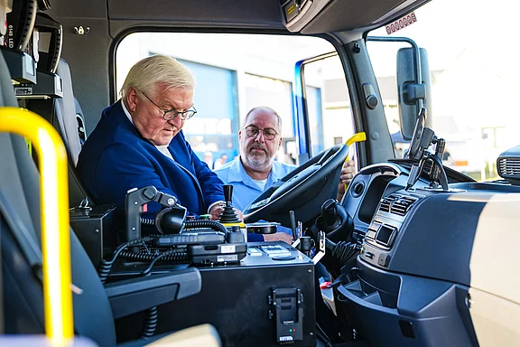Zwei Männer sitzen im Fahrersitz eines großen Fahrzeugs. Der Mann auf der linken Seite betrachtet die Bedienelemente, während der Mann auf der rechten Seite aufmerksam zuschaut. Beide wirken interessiert und sprechen miteinander.