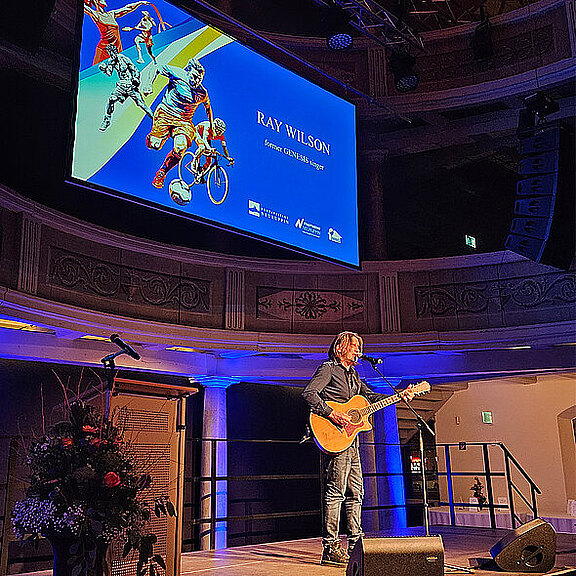 Ein Mann steht auf einer Bühne mit einer Gitarre. Er hat lange, hellbraune Haare und trägt ein schwarzes Hemd. Im Hintergrund ist ein Bildschirm mit dem Namen "Ray Wilson" und einem bunten grafischen Design, das Sportler zeigt. Es scheint eine Live-Darbietung zu sein.