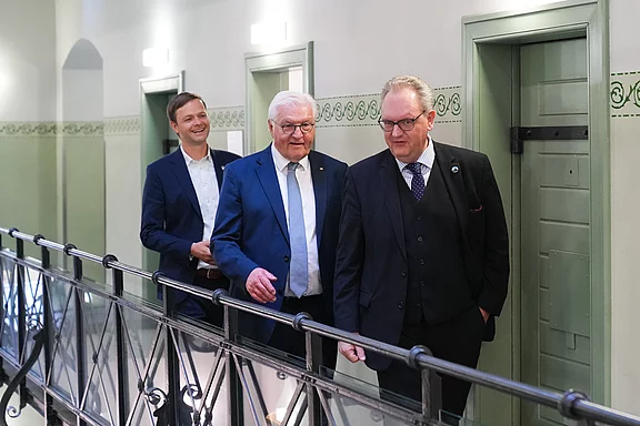 Drei Männer gehen in einem Flur. Der Mann ganz vorne trägt einen dunklen Anzug und eine Krawatte, während die beiden anderen ebenfalls formell gekleidet sind. Sie scheinen sich zu unterhalten und bewegen sich in Richtung einer Tür.