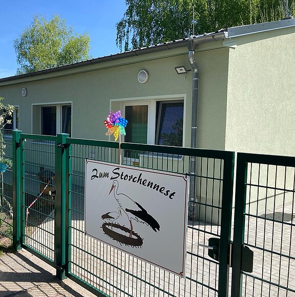 Eingangstor vor einem kleinen, hellgrünen Gebäude. Am Zaun hängt ein Schild mit der Aufschrift „Zum Storchennest“ und einer Storchgrafik. Daneben dreht sich ein buntes Windrad.