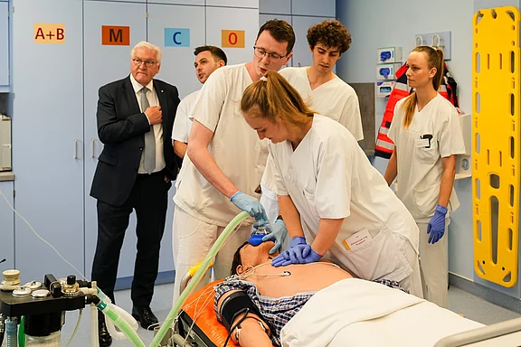 Eine Gruppe von medizinischem Personal führt eine Notfallbehandlung an einem Patienten durch, der auf einer Liege liegt. Eine Person bedient dabei ein Beatmungsgerät, während andere beobachten. Im Hintergrund steht ein Mann in Anzug, der das Geschehen interessiert verfolgt.