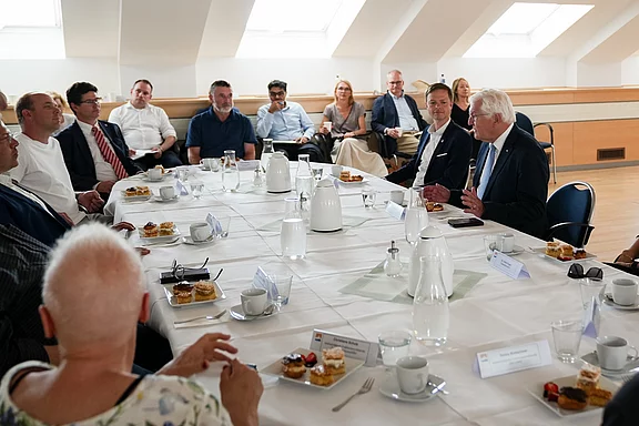 Ein Tisch ist mit Getränken und Snacks gedeckt. Eine Gruppe von Männern und Frauen, die in geschäftlicher Kleidung gekleidet sind, sitzt um den Tisch. Ein älterer Mann spricht, während andere aufmerksam zuhören. Es scheint sich um eine formelle Diskussion oder ein Meeting zu handeln.
