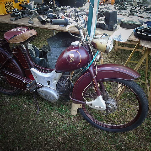 Ein antikes Motorrad in rot-brauner Farbe steht auf einer Wiese. Es hat einen klassischen Look mit einer runden Frontscheinwerfer, einem bequemen Sitz und technischen Teilen im Hintergrund, die auf einem Tisch ausgelegt sind.