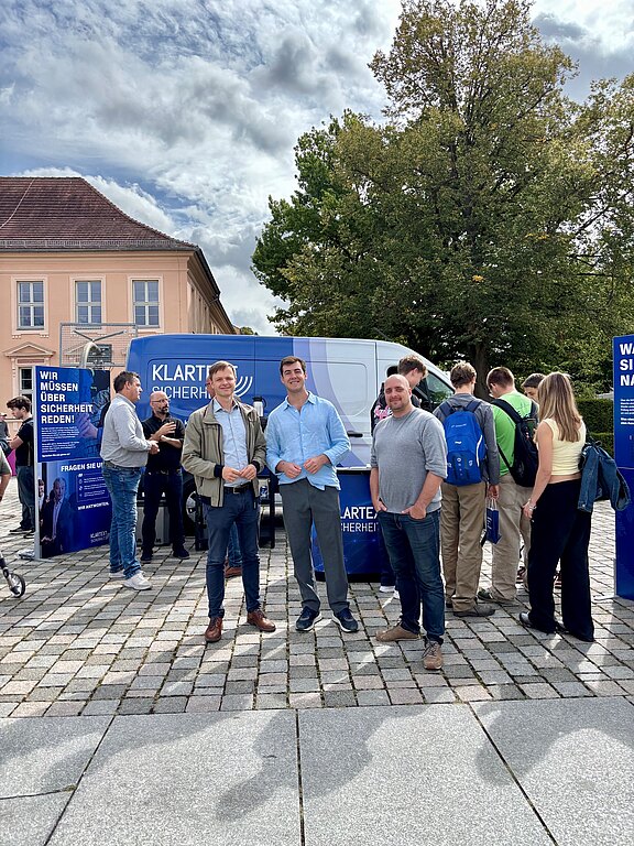Menschen versammeln sich auf einem Platz an einem blauen Infostand mit Van. Drei Männer stehen vorn, weitere Personen sprechen dahinter. Auf den Tafeln steht Klartext Sicherheit und Wir müssen über Sicherheit reden. Bäume und Gebäude im Hintergrund, bewölkter Himmel.