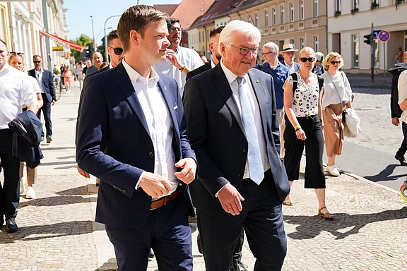 Der deutsche Bundespräsident und ein Begleiter gehen in einer Stadtstraße entlang. Sie tragen formelle Kleidung und scheinen ein Gespräch zu führen, während im Hintergrund weitere Personen und Gebäude sichtbar sind. Es ist ein sonniger Tag.