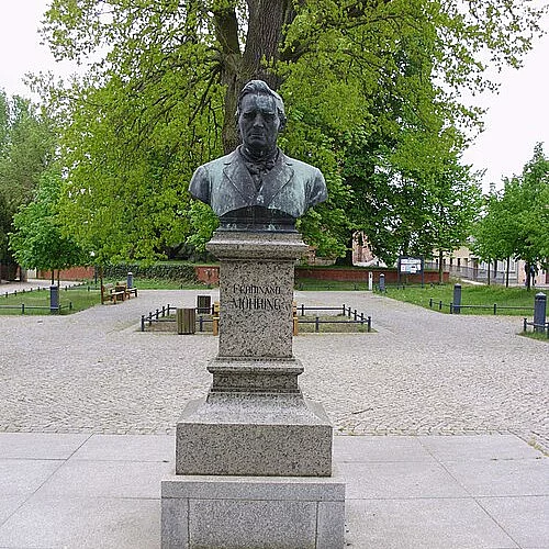 Bronzene Büste eines Mannes auf hohem Granitsockel, zentral auf einem gepflasterten Platz. Im Hintergrund Bäume und Bänke. Das Denkmal steht im Mittelpunkt der Szenerie.