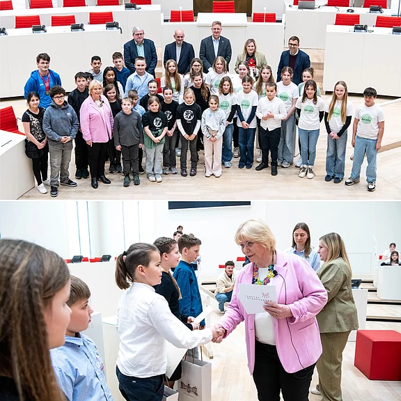 In einem modernen Sitzungssaal posiert eine große Gruppe Schulkinder mit mehreren Erwachsenen für ein Foto. Darunter: eine Frau überreicht Schülern eine Urkunde und Tüte, gibt einem Kind die Hand. Atmosphäre: offizielle Veranstaltung oder Auszeichnung.