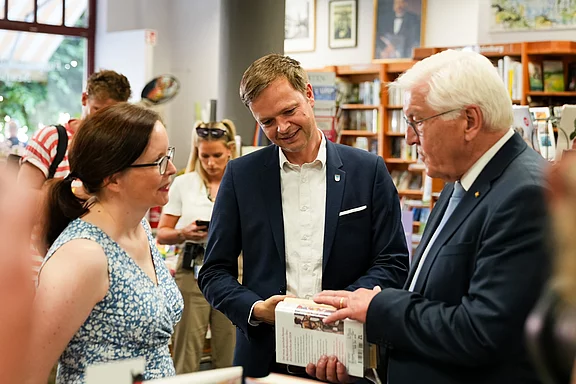 Drei Personen stehen in einer Buchhandlung. Eine Frau mit Brille trägt ein gemustertes Kleid und spricht mit zwei Männern, von denen einer ein Buch in der Hand hält. Alle scheinen in ein Gespräch vertieft und zeigen Interesse an dem Austausch.