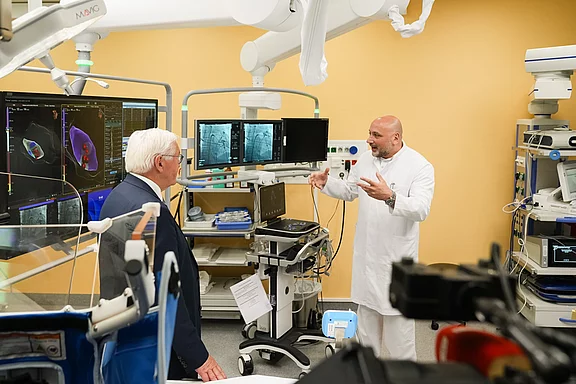 Ein Mediziner erklärt einem Zuhörer in einem modernen Operationssaal die Technologie, die auf Bildschirmen angezeigt wird. Im Hintergrund sind medizinische Geräte und Bildschirme sichtbar, die verschiedene Daten und Bilder zeigen.