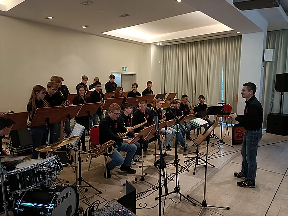Eine Jugendbigband spielt in einem großen Raum. Der Leiter steht vorne und dirigiert. Die Musiker sind auf Stühlen verteilt, jeder mit einem Notenständer. Instrumente wie Saxophone und Trompeten sind gut sichtbar, während ein Schlagzeug auf der linken Seite steht.
