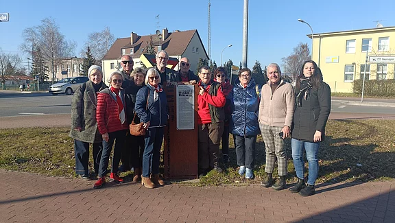 Eine Gruppe von 14 Personen steht um ein Info-Schild herum, das auf einem Bürgersteig aufgestellt ist. Die Personen sind in unterschiedlichen Winterjacken gekleidet und lächeln in die Kamera. Im Hintergrund sind einige Gebäude und Bäume sichtbar. Es ist ein klarer, sonniger Tag.