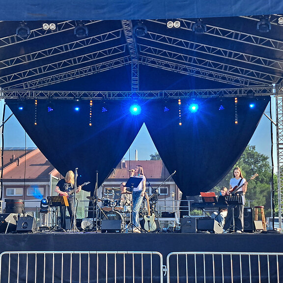 Eine Band steht auf einer Bühne mit einem dunklen Vorhang. Strahlende Lichter beleuchten die Musiker, die verschiedene Instrumente spielen. Im Hintergrund sind weitere Bandmitglieder erkennbar. Die Atmosphäre ist lebhaft und musikalisch.