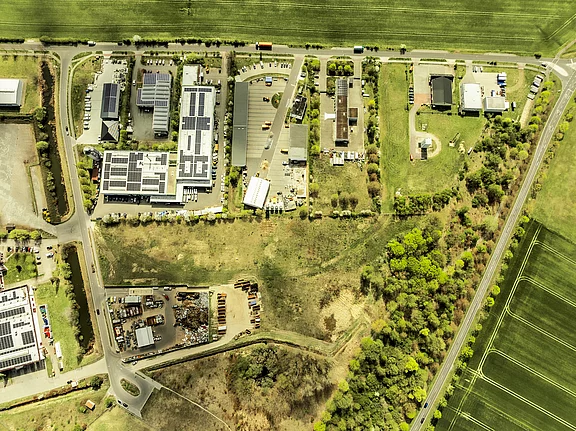 Senkrechte Luftaufnahme eines Gewerbegebiets zwischen Feldern: mehrere Hallen mit Photovoltaikanlagen auf den Dächern, Parkplätze und Zufahrtsstraßen. Rechts Wald- und Wiesenflächen, links unten ein offenes Materiallager. Umgebende Landstraßen rahmen das Areal.