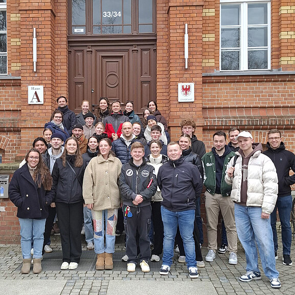 Eine Gruppe von etwa 30 Personen steht vor einem Gebäude mit Ziegelmauern. Die Menschen sind in verschiedenen Kleidungsstilen gekleidet und lächeln in die Kamera. Es scheint sich um eine Gemeinschaft oder ein Gruppenevent zu handeln.