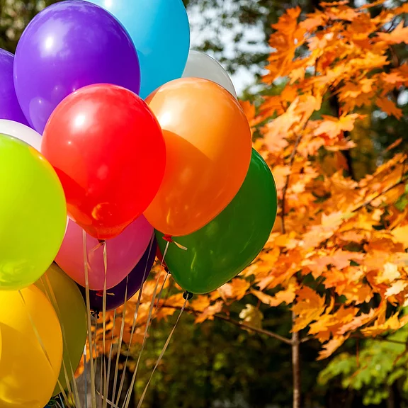 Bunte Heliumballons in Rot, Orange, Lila, Grün und Gelb schweben draußen; im Hintergrund leuchten orangefarbene Herbstblätter.