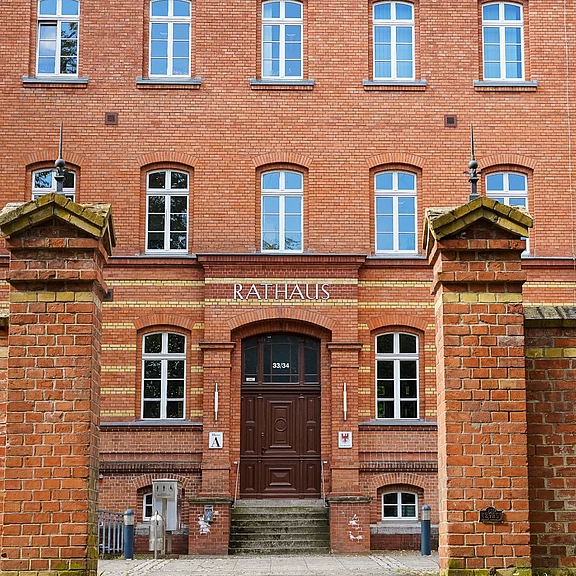 Das Bild zeigt das Rathaus eines Stadtteils, mit einer rotbacksteinernen Fassade. Der Eingang ist in der Mitte des Gebäudes und trägt über der Tür die Aufschrift "RATHAUS". Vor dem Gebäude befinden sich zwei Mauertürme, die den Eingang flankieren.
