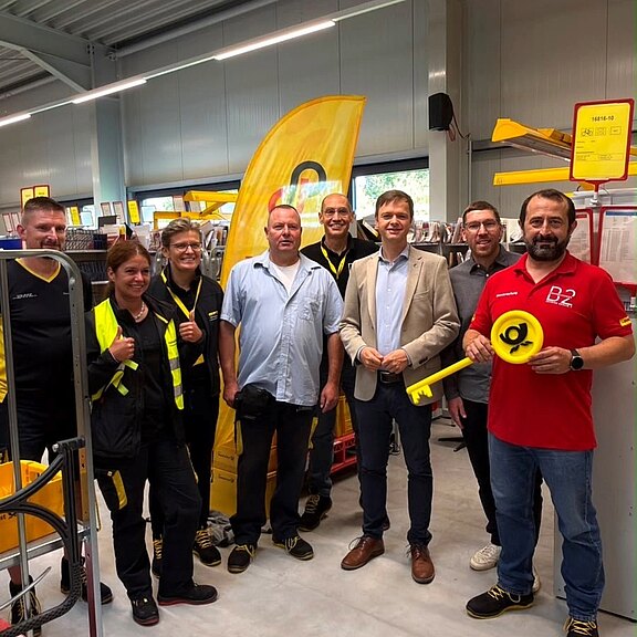 Gruppe von acht Personen in einem Postverteilzentrum, mehrere in gelben Uniformen, posieren für ein Foto. Ein Mann im roten Shirt hält einen großen gelben symbolischen Schlüssel. Regale und Postkisten im Hintergrund. Stimmung freundlich.