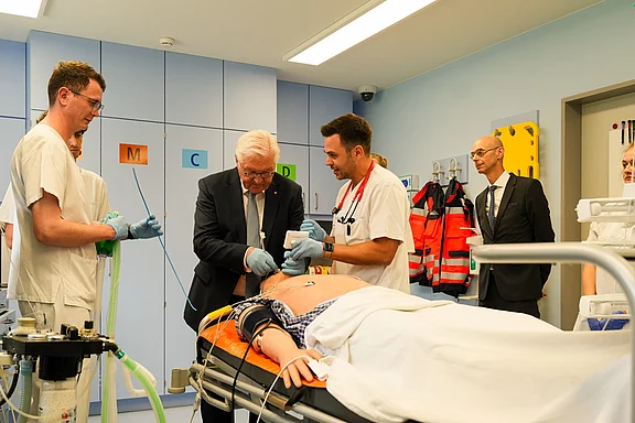 Auf dem Bild sind mehrere Personen in einem Krankenhauszimmer zu sehen. Zwei Ärzte behandeln einen Patient, während eine weitere Person assistiert. Im Hintergrund stehen Zuschauer, die den Prozess beobachten. Die Szene vermittelt einen Eindruck von medizinischer Notfallversorgung.